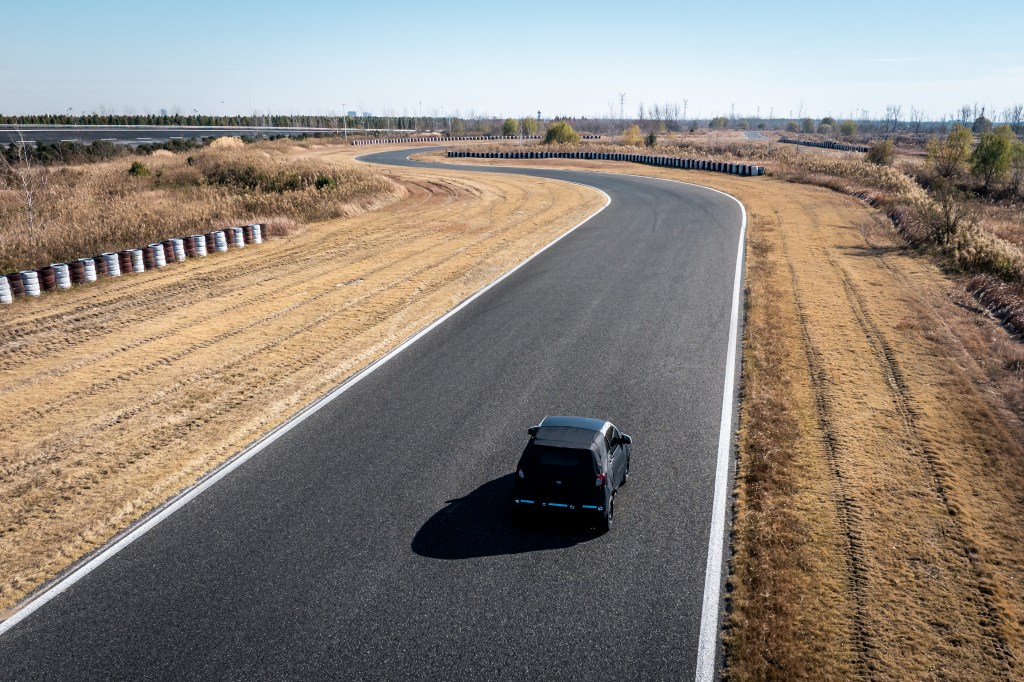 A camouflaged Smart #2 prototype is seen from above driving on a curved asphalt test track, with dry grass and red-and-white barriers lining the circuit.